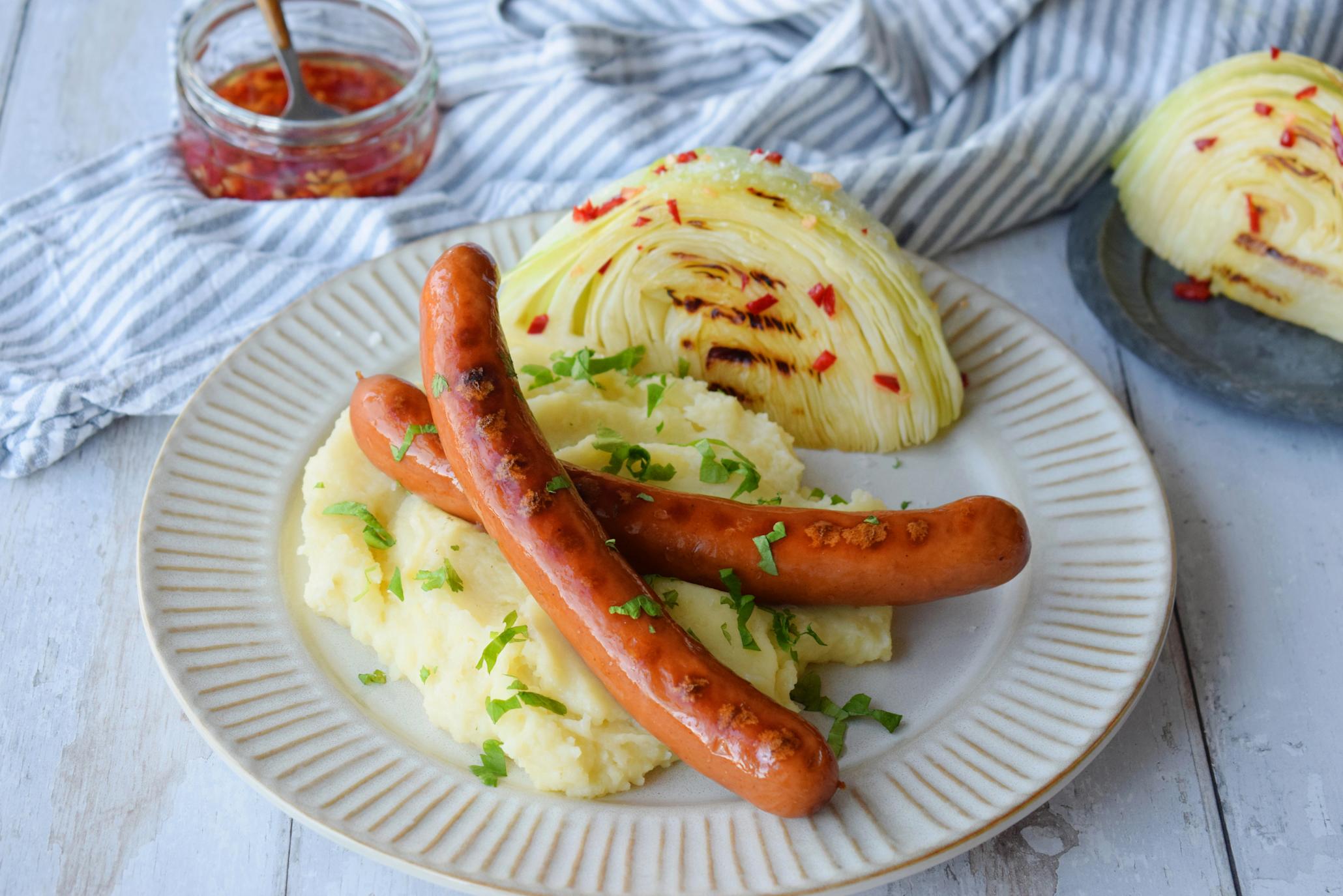 To stykk Cheesy bacon pølse, servert over potetmos på lys tallerken. Bladpersille strødd over, og grillet nykål på siden av tallerkenen, med grillolje av chili og hvitløk.