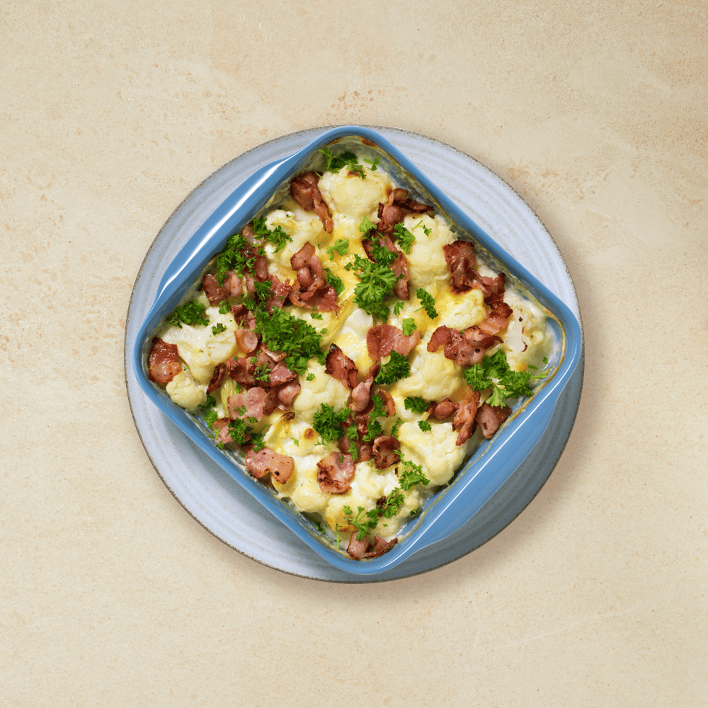 Gratinert blomkål med bacon, servert i en blå ildfast form som står på en grå tallerken på beige bakgrunn. Friske urter strødd over.