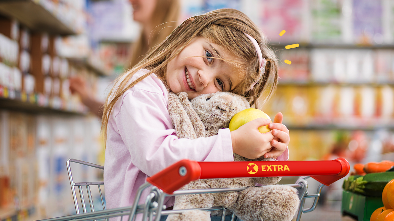 En jente sitter i en Extra handlevogn i en butikk mens hun holder et eple og en bamse. 