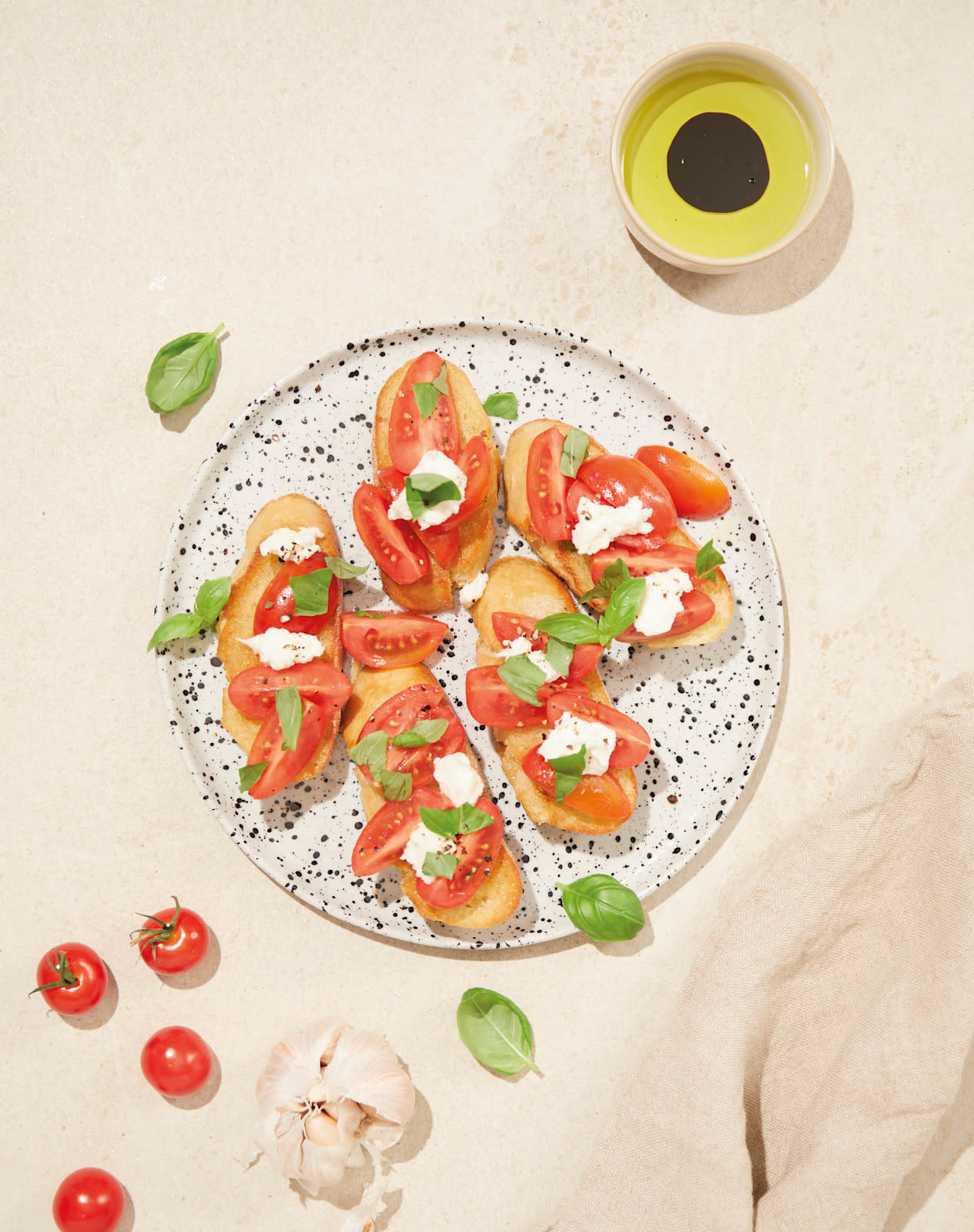 Bruschetta med tomat og mozzarella servert på lyst prikkete fat med frisk basilikum og nykvernet pepper. Liten skål olivenolje og balsamiko på siden.