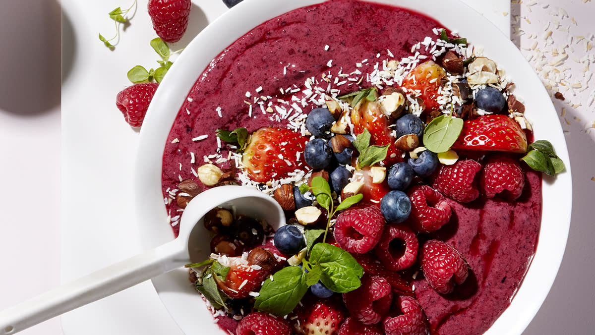 Smoothiebowl med friske bær og ristede nøtter, i hvit skål, toppet med kokosmasse, nøtter, bringebær, jordbær, blåbær og frisk mynte.