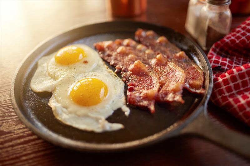 To speilegg med tre skiver bacon i svart støpejernspanne, på trebord. Rødrutet kjøkkenhåndkle.