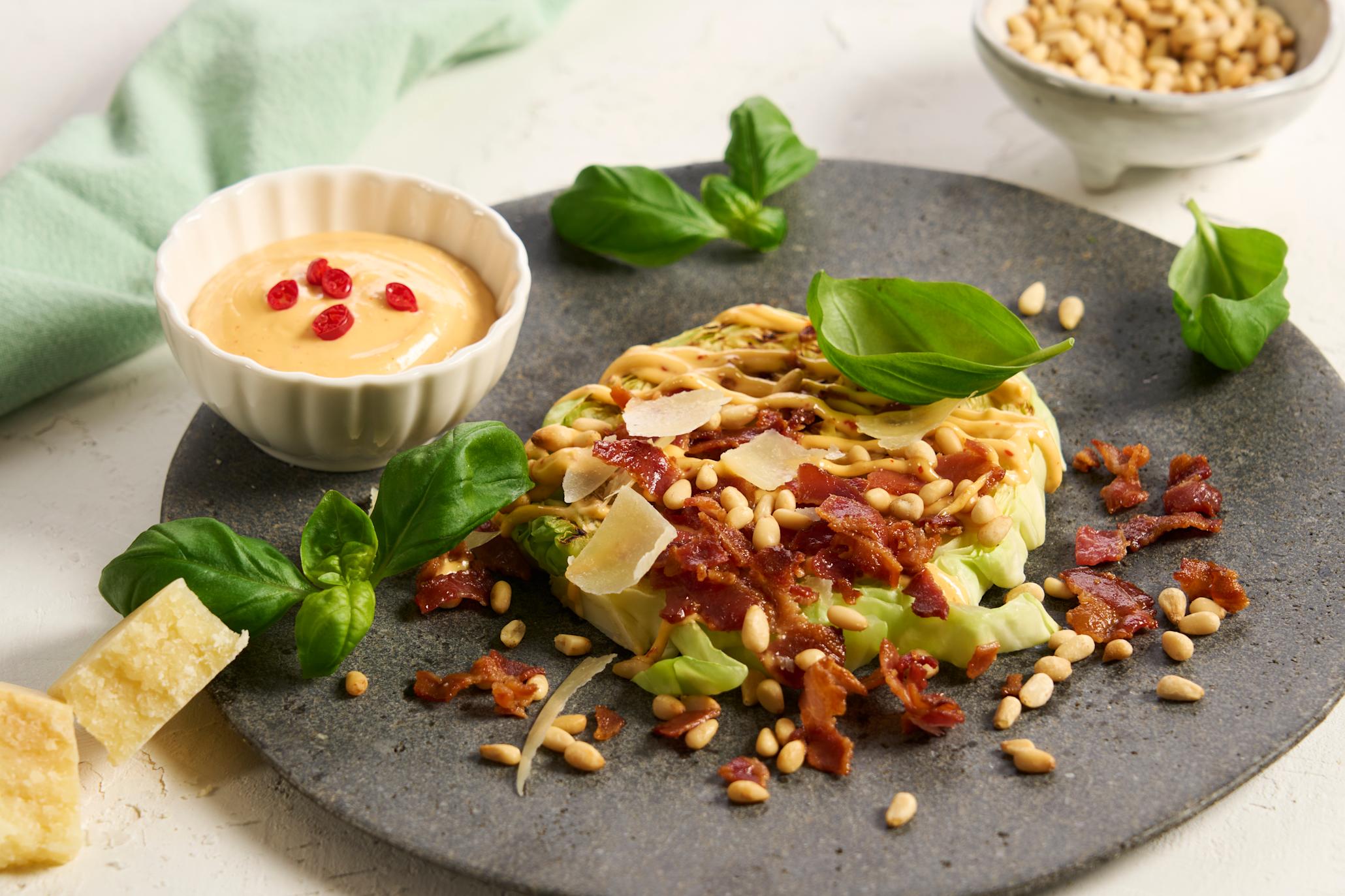 Nykålbiff med parmesan, bacon og aioli med chili servert på rund skiferhelle med friske basilikumblader og pinjekjerner. Små skåler med chiliaioli og pinjekjerner ved siden av.