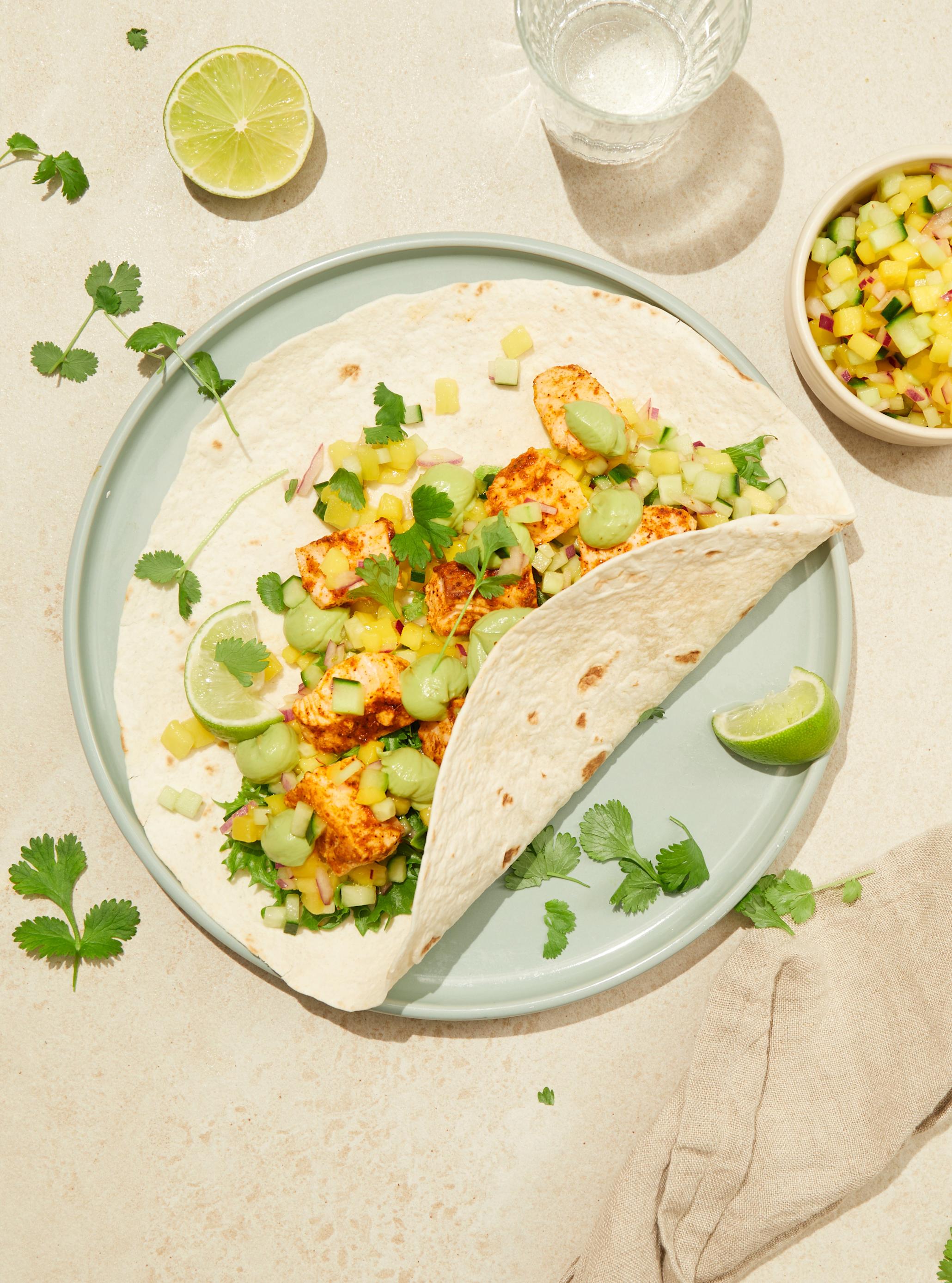Fisketaco med hjemmelaget guacamole og mangosalsa servert i tacolefse på lyseblå tallerken, med frisk koriander og limebåter til. Liten skål med mangosalsa, og vannglass ved siden av.