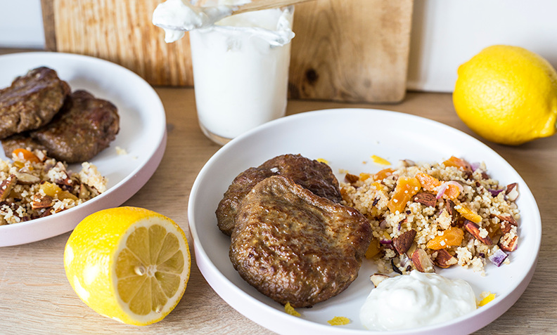 Karbonader servert på hvit middagstallerken, på trebord. Fetadressing, og couscous med aprikos, mandler og rødløk ved siden av. Ferske sitroner og glass med fetadressing i bakgrunnen.