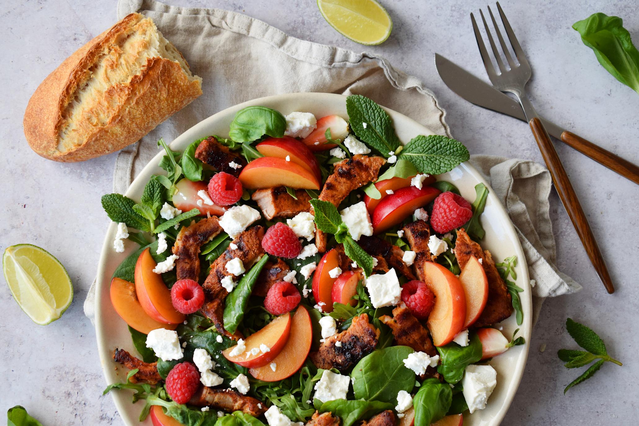 Sommersalat med grillribbe, fetaost, plommer, bringebær, jordbær, babyleaf, ruccola og ferske urter, på lys tallerken. Lys bakgrunn med limebåter, baguette og ferske urter.