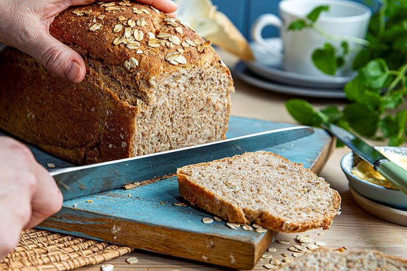 Hjemmelaget brød bakt med havregrøt, skjæres på blå trefjøl. Brødet har et dryss av havregryn, og i bakgrunnen er det ferske urter, og kaffekopper.