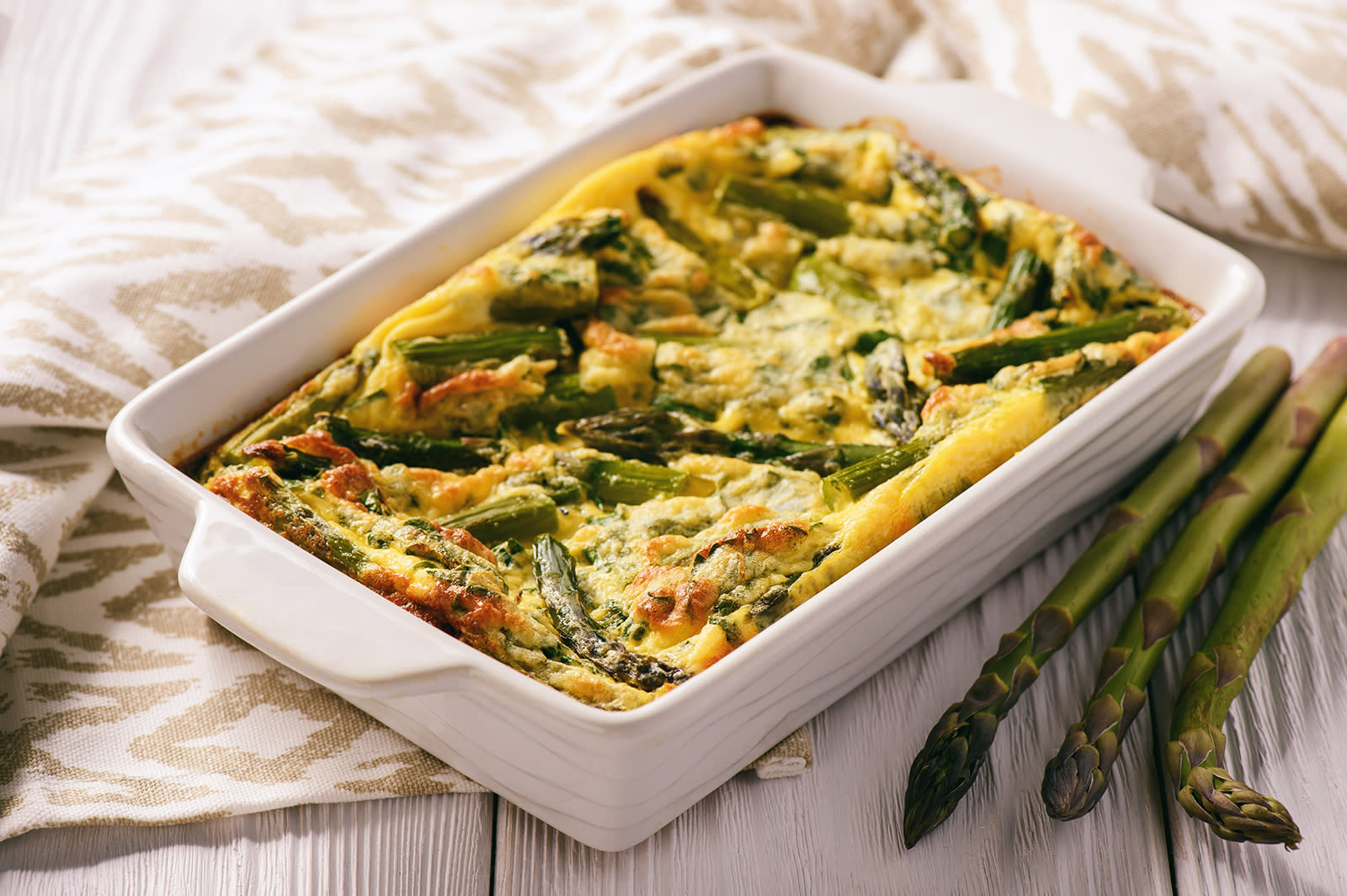 Frittata med asparges servert i firkantet hvit ildfast form på lyst trebord med mønstret kjøkkenhåndkle ved siden av.