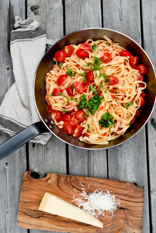 Kokt tagliatelle med ferske tomater, basilikum og parmesan presentert i jernpanne på trebakgrunn, med en bit parmesan ved siden av.