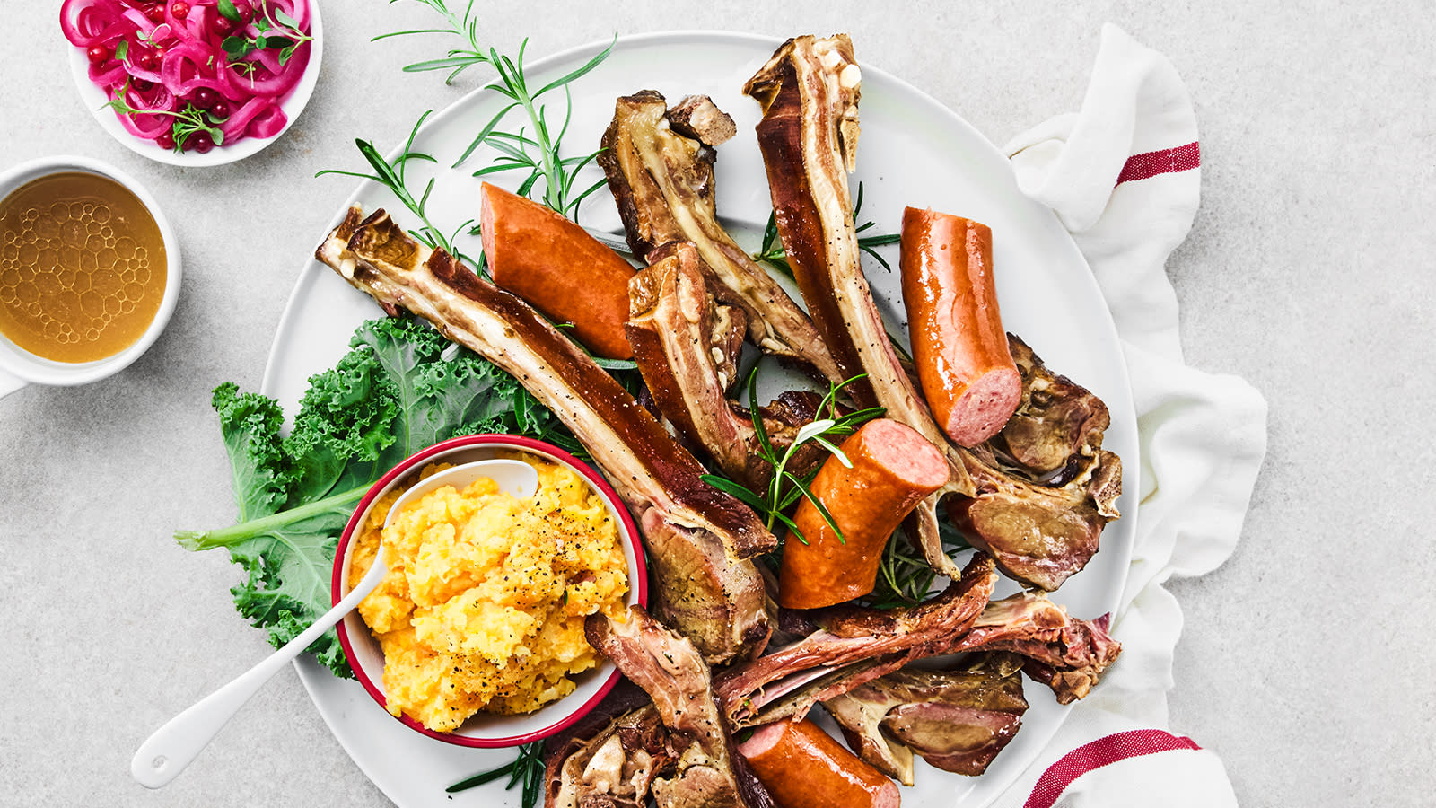 Pinnekjøtt i grønnsakskraft med rotmos og syltet rødløk servert på hvit tallerken med rotmos, vossakorv og frisk rosmarin. Små skåler med grønnsakskraft og syltet rødløk ved siden av.