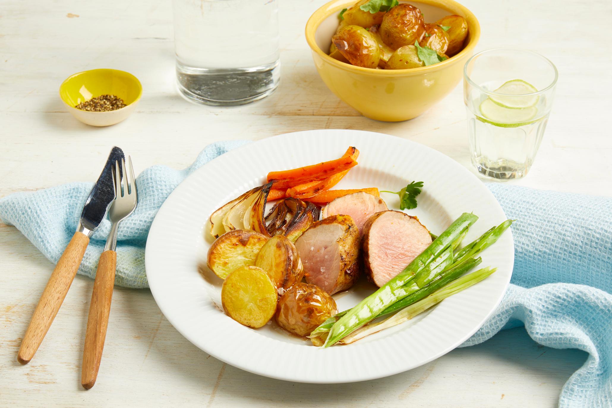 Indrefilet av svin med appelsin- og hvitløksmarinade og ovnsbakte grønnsaker servert i skiver på hvit tallerken på lyst dekket middagsbord. Gul skål med ovnsbakte poteter i bakgrunnen.