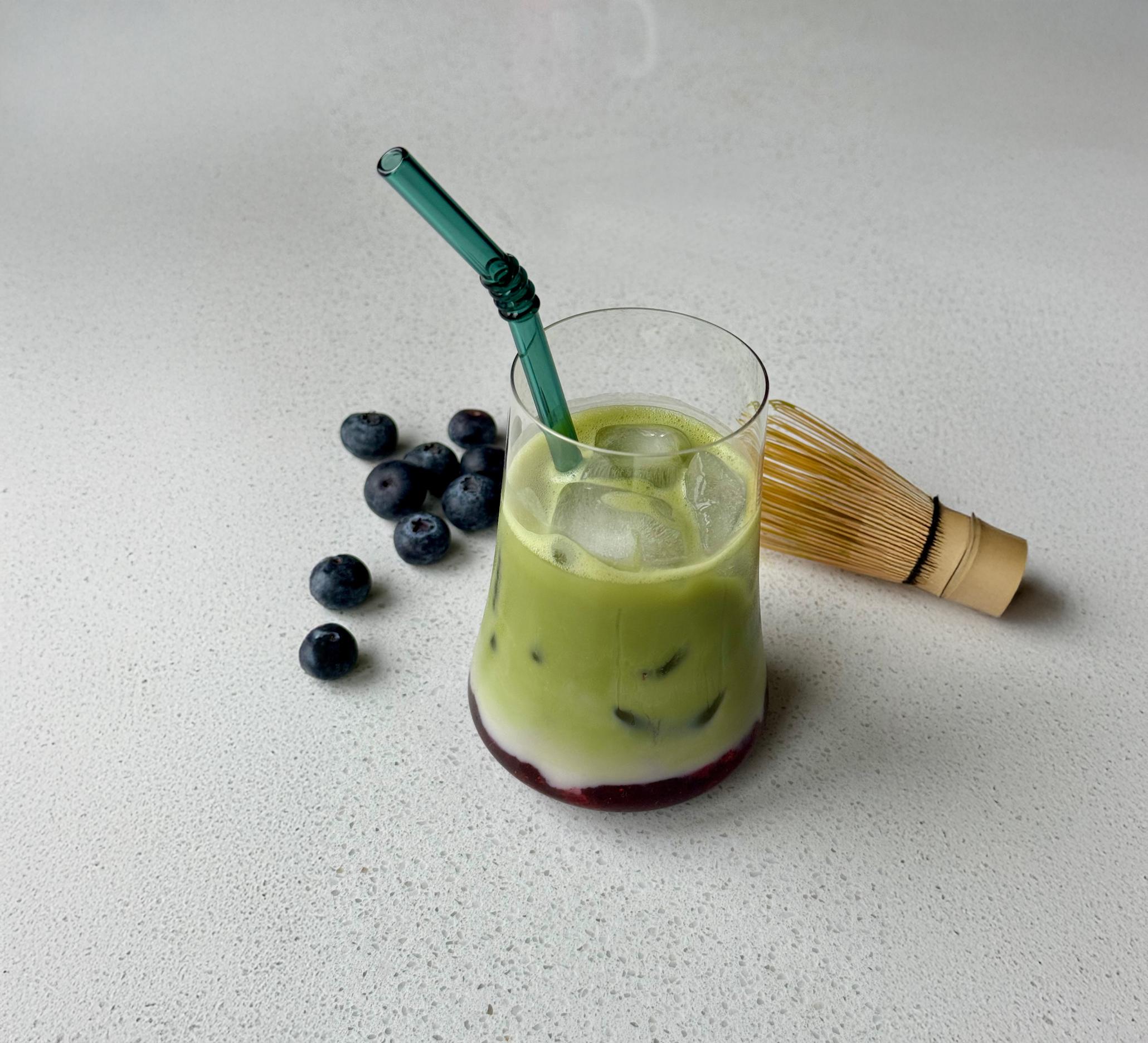 Matcha islatte med blåbærsirup servert i et glass med isbiter og grønt sugerør av glass. Matchavisp og ferske blåbær ved siden av.
