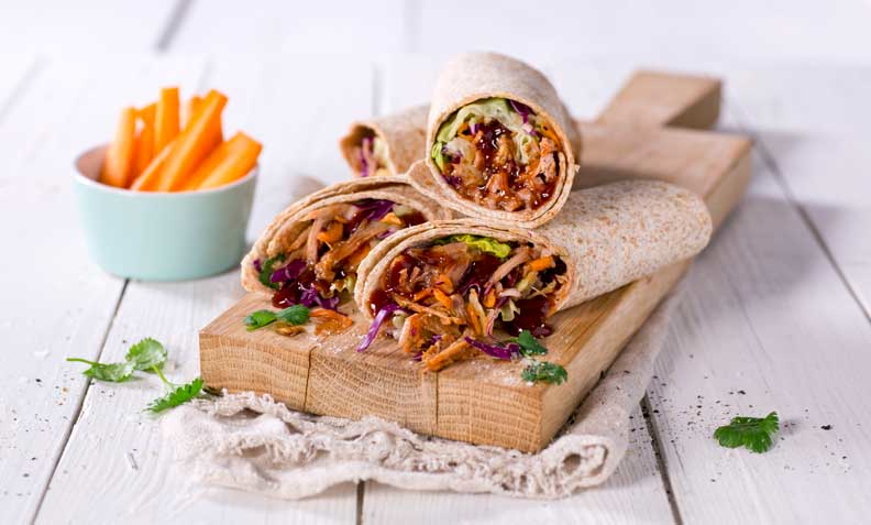 Wrap med pulled pork, råkostsalat og BBQ glaze på trefjøl. Hvit trebakgrunn. Lyseblå skål med gulrotstaver i bakgrunnen.