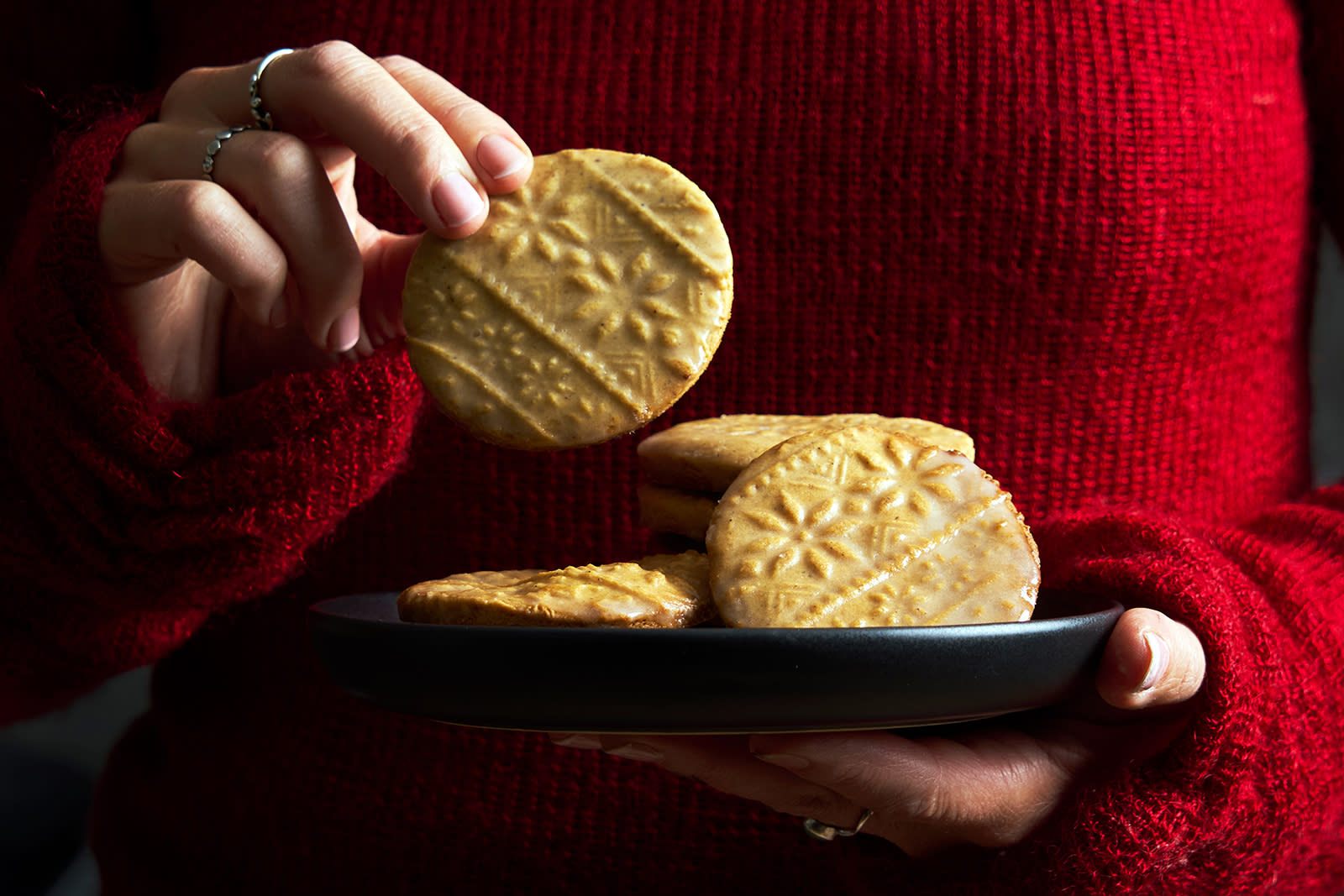 Pepperkakecookies med vaniljeglasur servert på fat holdes av kvinne med rød strikkegenser.