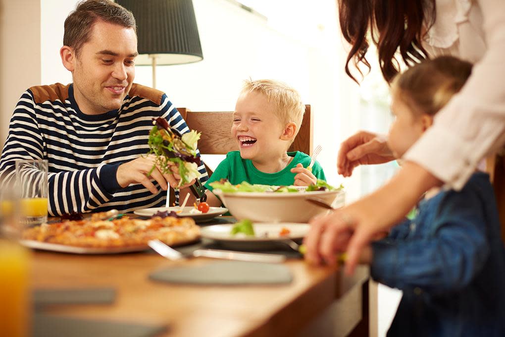 Situasjonsbilde av familie med små barn som sitter ved middagsbordet og spiser salat og pizza.