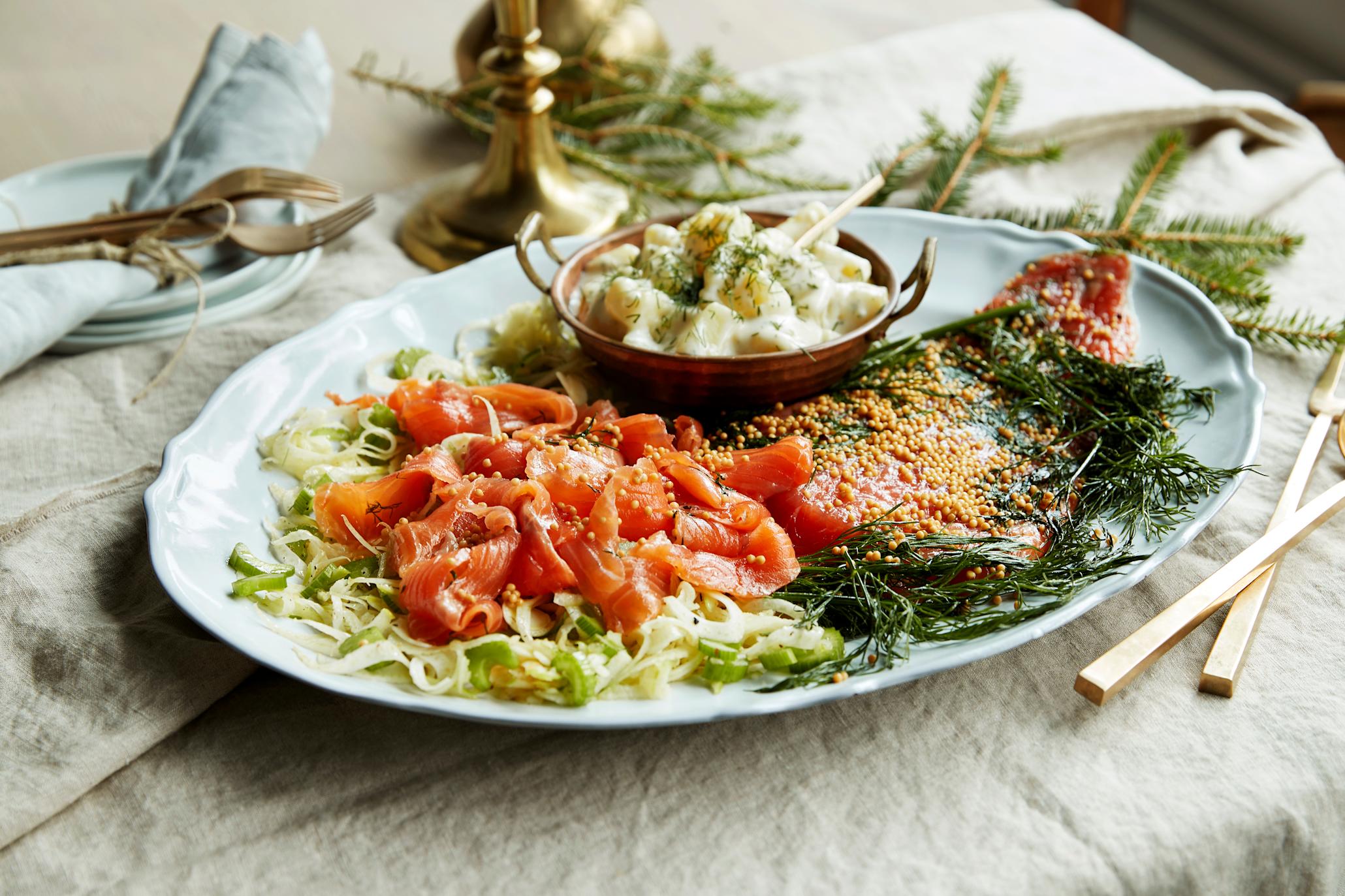Gravet ørret servert på avlangt hvitt fat på en seng av fennikelsalat og fersk dill. Dillstuede poteter i skål på fatet sammen med ørreten. Fatet står på bord med linduk, granbar og gullfarget bestikk.