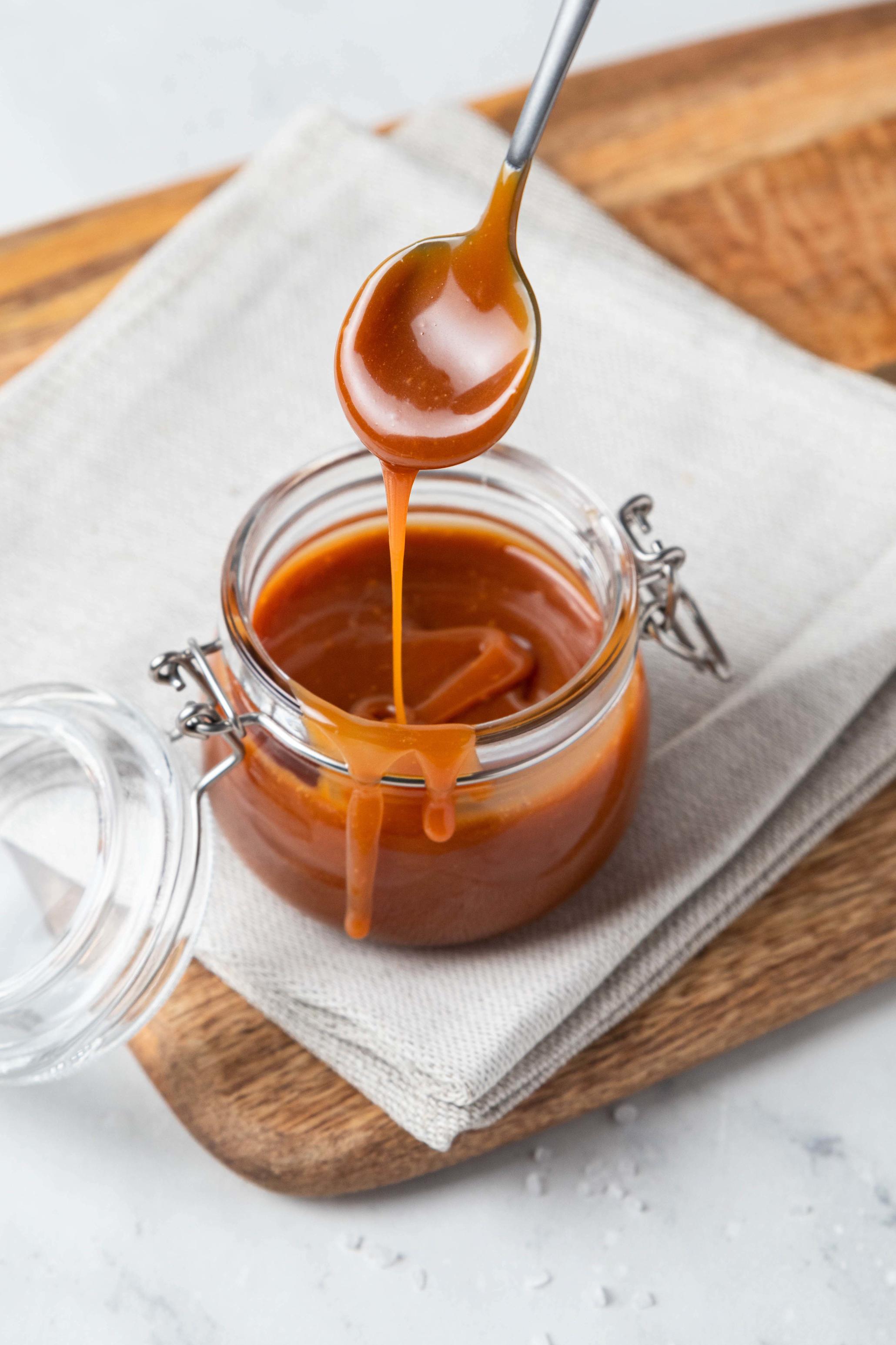 Hjemmelaget karamellsaus til iskrem, lagt på syltetøyglass, som står på trefjøl med linserviett. En skje løfter karamellsaus ut av glasset.