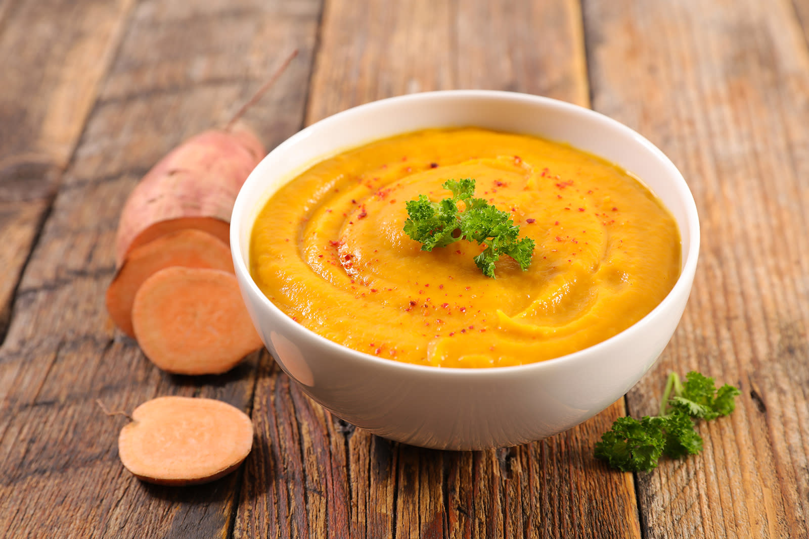 Tykk søtpotetsuppe i skål, pyntet med kruspersille, med en oppskåret rå søtpotet ved siden av, servert i hvit bolle på rustikk trebord.
