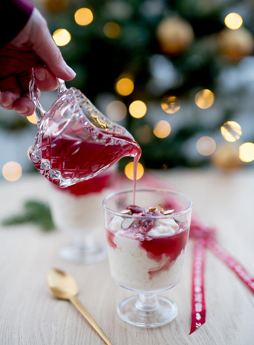 Ris a la mande på glass, står på lyst trebord, med juletre i bakgrunnen. Hånd holder en liten glassmugge som heller rød saus over dessertglasset.