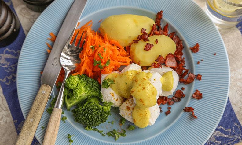 Fiskeboller i hvit saus med karri, kokte poteter og sprøstekt bacon på lys blå tallerken. Revet gulrot og kokt brokkoli ved siden av.