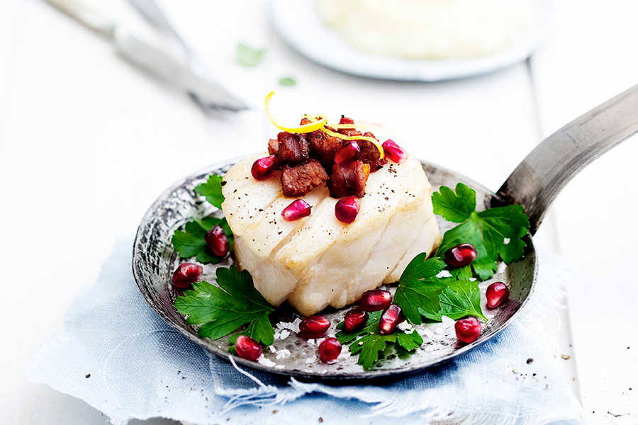Skrei servert på en liten metallpanne med ferske urter, granateplefrø og baconbiter på toppen. Pannen står på blått tøystykke på et hvitt bord, med bestill i bakgrunnen.