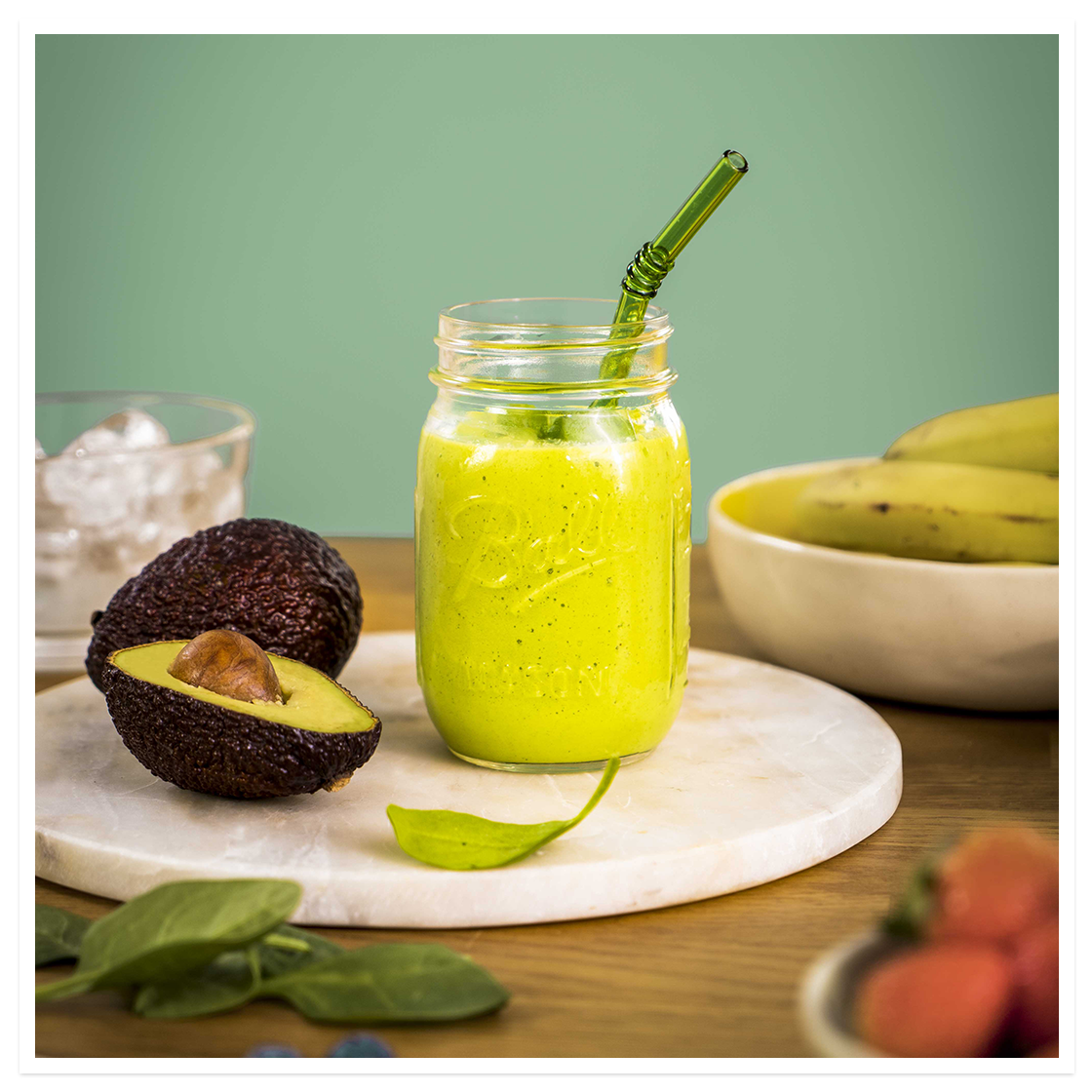 Sommergrønn smoothie med avokado, banan og spinat, i glass på hvit marmorfjøl med avokado, banan og spinat rundt.