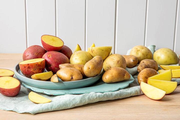 Forskjellige sorter poteter i gult, rødt og brunt, både hele og delt, ligger på lys blå tallerken og lys blå trefjøl. 