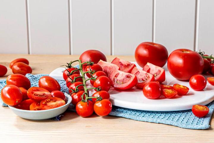 Tomater av forskjellige slag ligger på fat, i skål og på et trebord. Noen av tomatene er skåret opp, andre er hele, og andre på klase. Blått kjøkkenhåndkle.