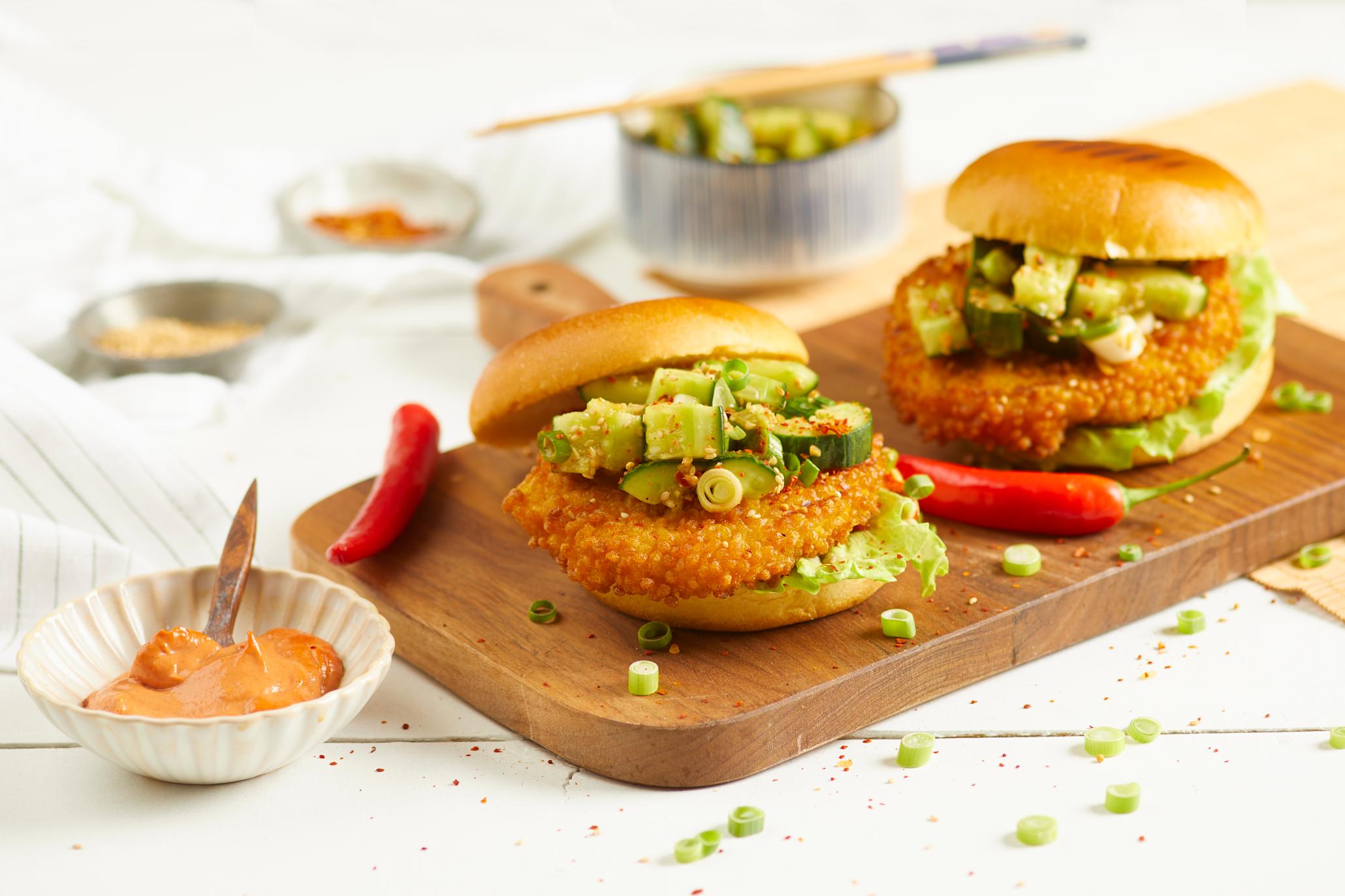 Grill Perfekt Crispy kyllingburger med asiatisk knust agurk salat og spicy gochujangmajo servert på mørk trefjøl på hvitt trebord. Liten hvit skål med gochujangmayo på siden, og asiatisk skål med agurksalat i bakgrunnen.