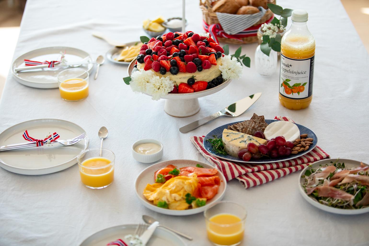 17. mai festdekket frokostbord med juice i glass. På bordet er det kake, eggerøre, røkelaks og et fat med oster og spekeskinke.