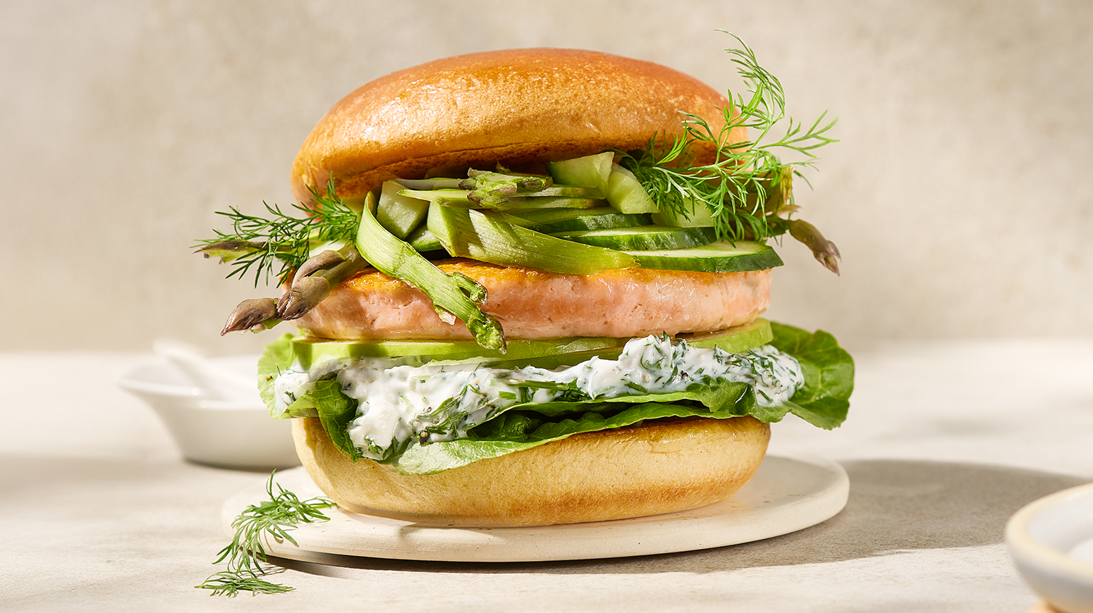 Lofoten lakseburger med frisk dillkrem og grønn crunch servert med høvlet asparges, agurk og frisk dill på beige asjett på beige bakgrunn.