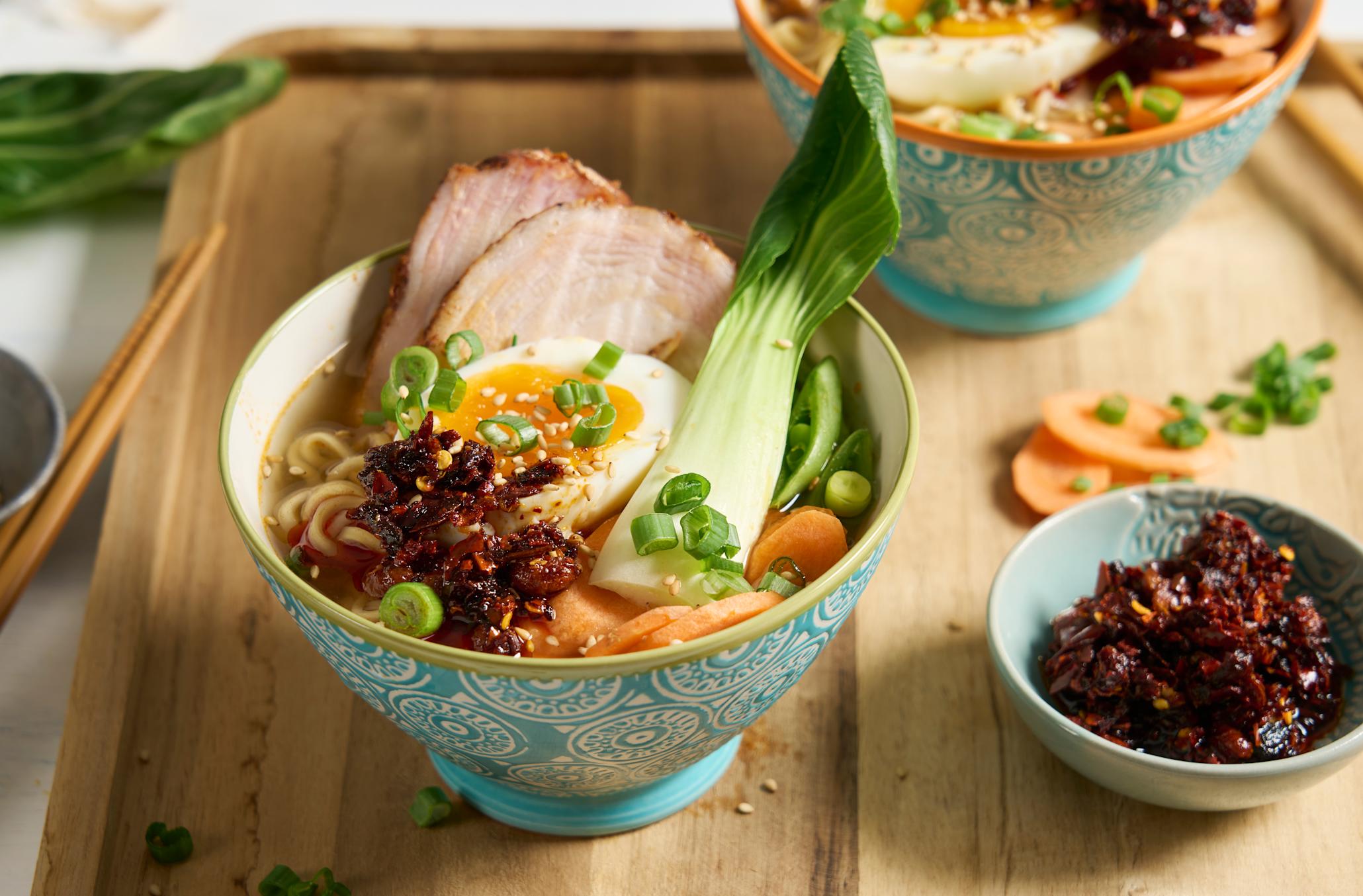 Pork belly ramen med bløtkokt egg og chili crisp servert i turkis skål med gulrot, pak choi, sesamfrø og vårløk. Skålen står på trefjøl med en annen skål med ramen. Liten skål med chilicrisp ved siden av.