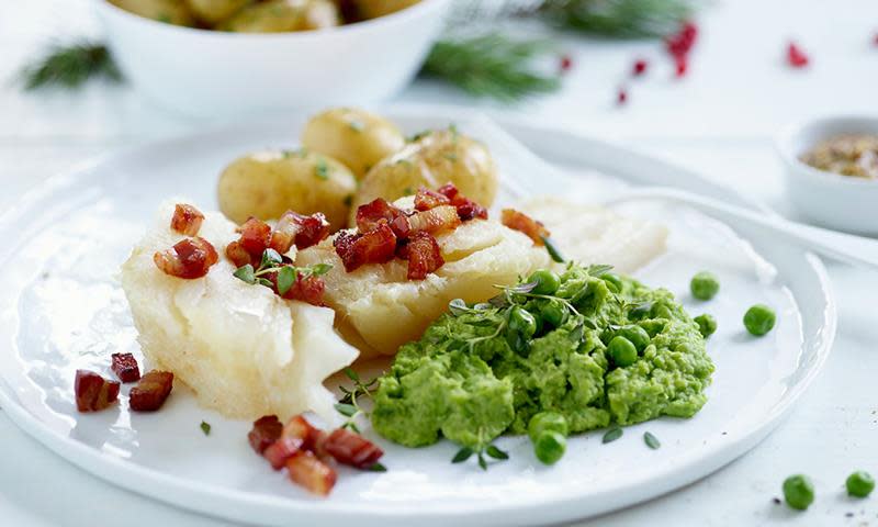 Hvit tallerken med lutefisk, kokte poteter, stekt bacon og perfekt ertepure til lutefisk. Tallerkenen står på hvit duk, med grønne og røde jule-elementer i bakgrunnen.