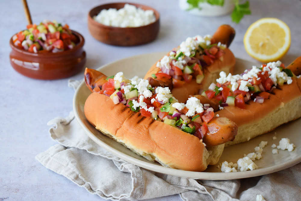 Tasty Garlic pølse med gresk salsa og fetaost, servert i pølsebrød på beige tallerken. Skåler med gresk salsa og fetaost i bakgrunnen.