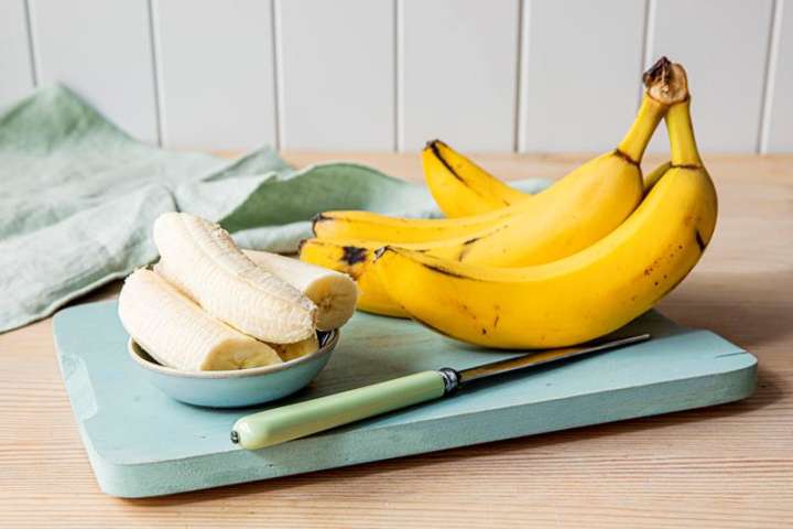Klase med bananer på blå skjærefjøl, ved siden av en skål med skrelte bananbiter. 