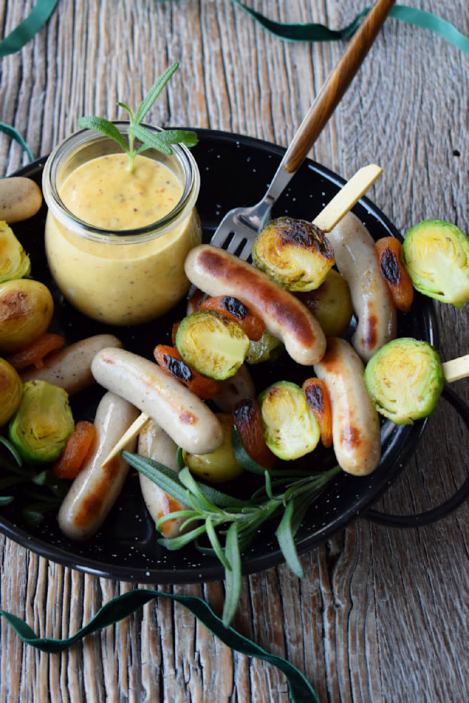 Sosisser på spyd med rosenkål, tørket aprikos og småpoteter servert i en sort rund form, med et glass honey mustard dip på siden. Formen står på et rustikk trebord.
