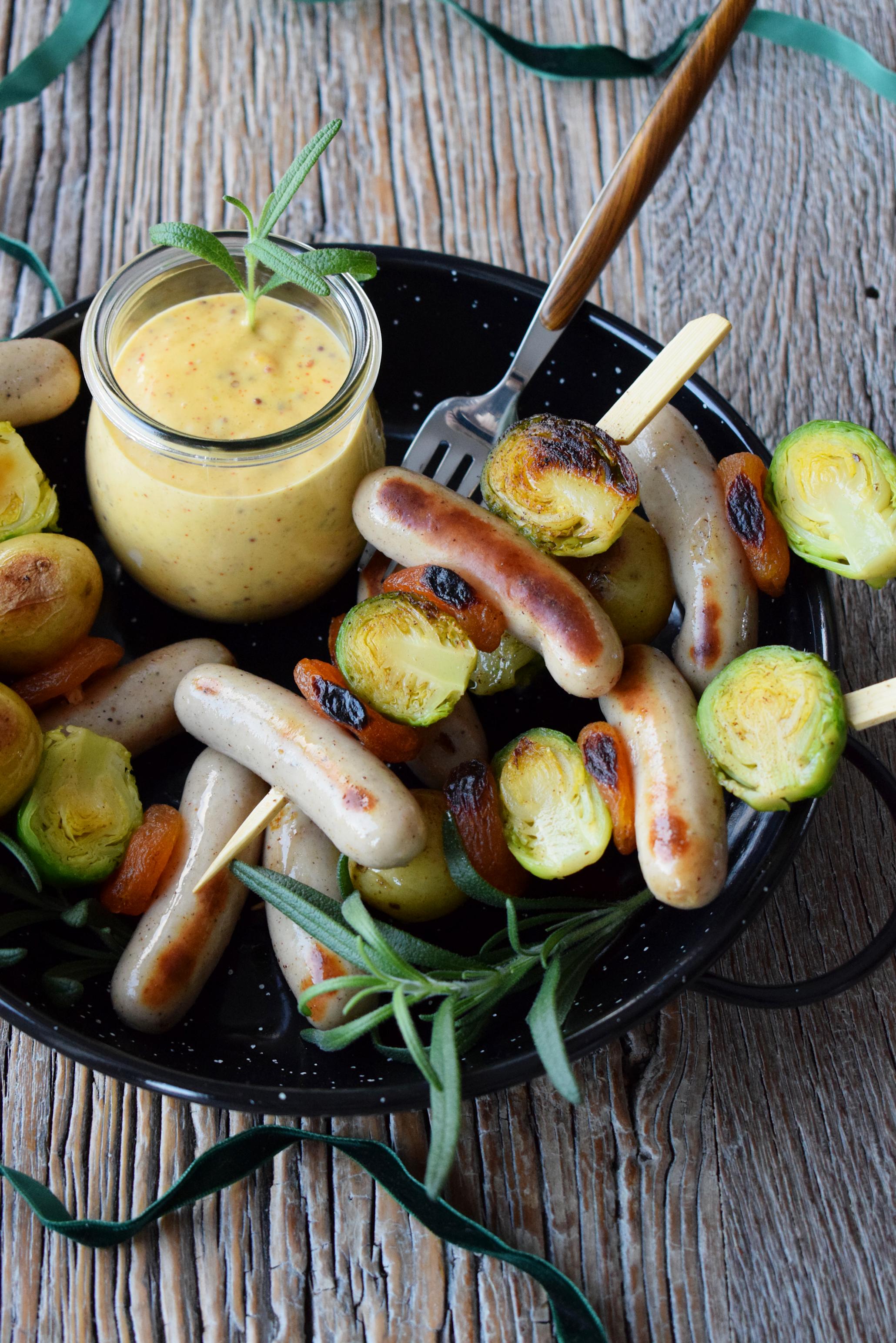 Coop sosisser på spyd med rosenkål, tørket aprikos og småpoteter ligger i en sort rund form, med et glass honey mustard dip på siden. Rustikk trebakgrunn med grønt julebånd.