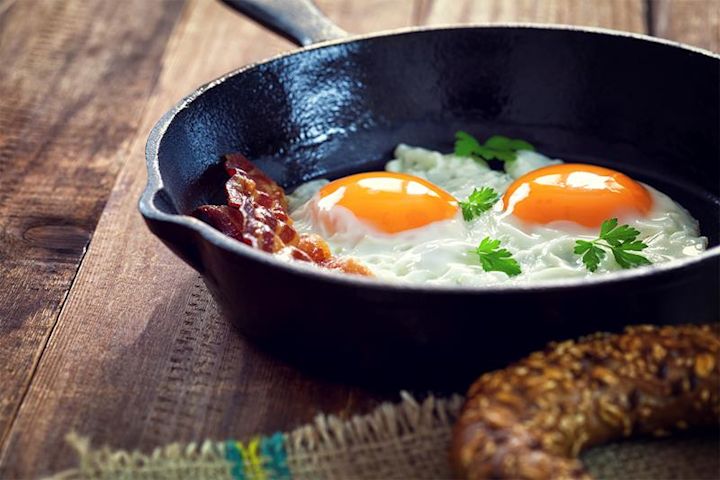 To speilegg i sort støpejernspanne med sprøstekt bacon og et dryss bladpersille. Rustikk trebakgrunn.