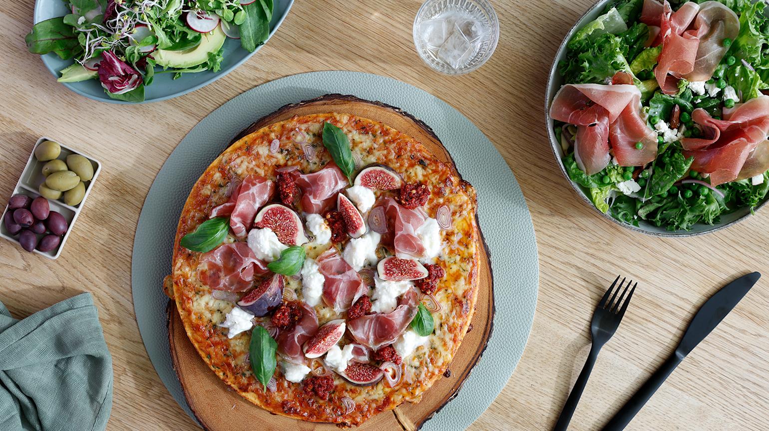 Pizza med skinke, fiken og mozzarella servert med to salater, oliven og et glass vann på et trebord.