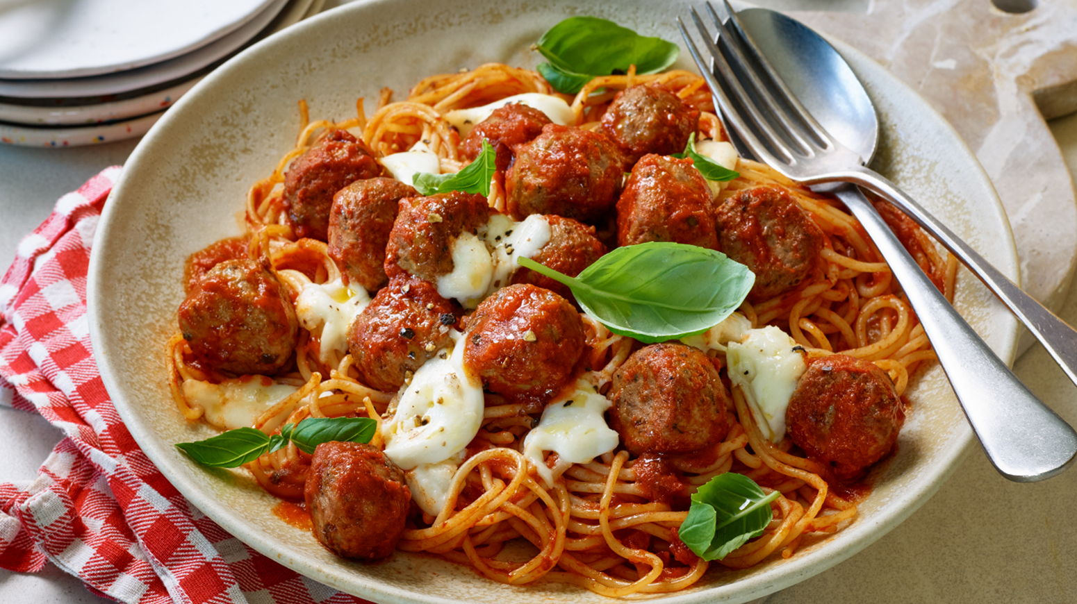 Tallerken med spaghetti og kjøttboller i tomatsaus, toppet med smeltet mozzarella og friske basilikumblader, servert med gaffel og skje på siden.