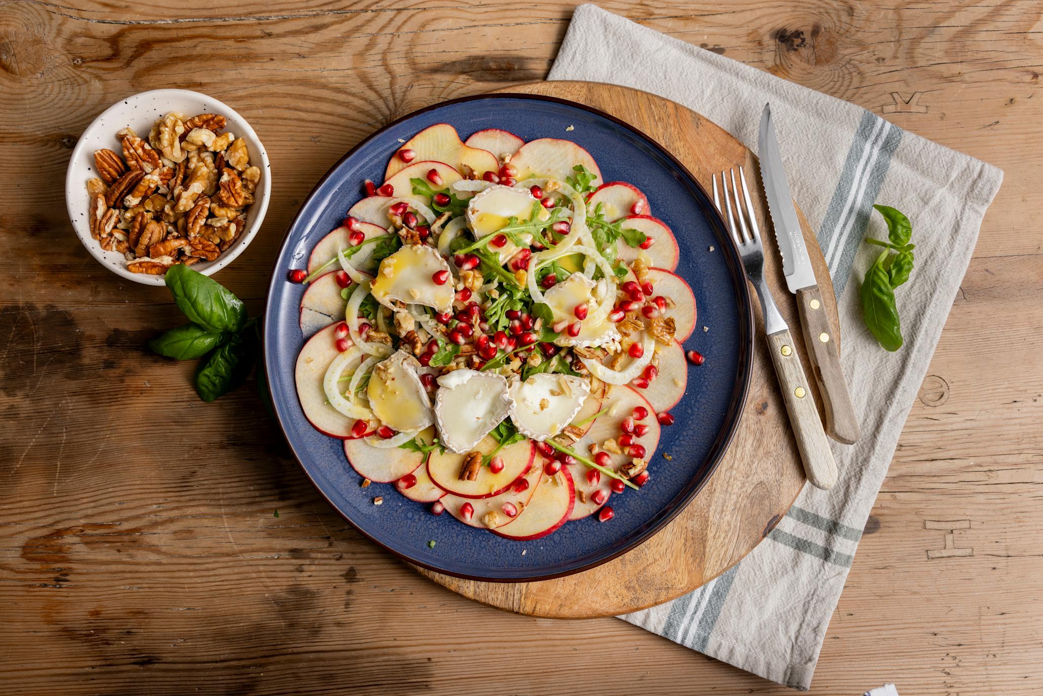 Carpaccio av eple og fennikel med chèvre, granateple og valnøtter servert på en blå tallerken på en trefjøl med bestikk og et kjøkkenhåndkle under. Liten skål med blandede nøtter ved siden av.