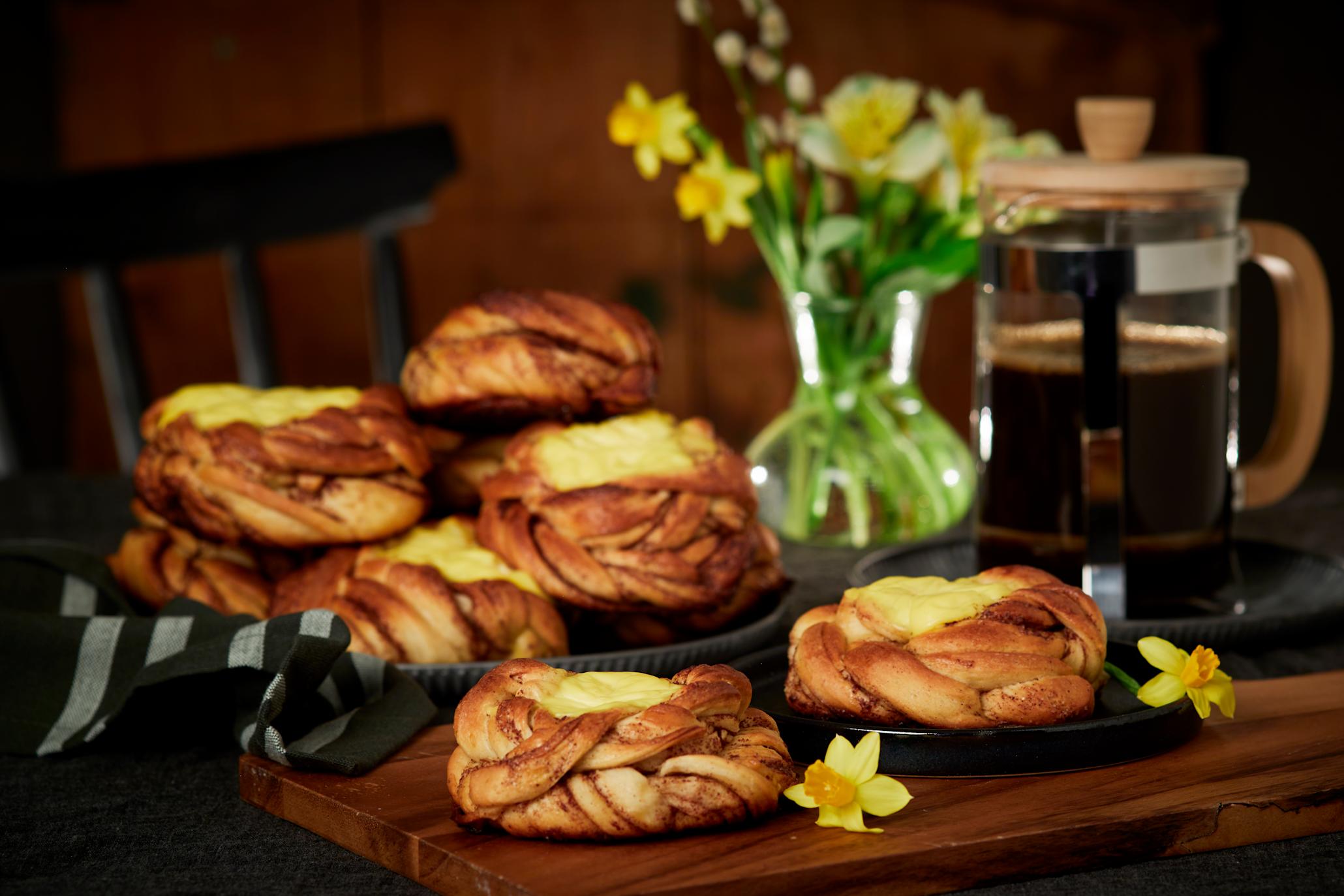 Kanelboller med vaniljekrem servert på både mørk tallerken og mørk trefjøl. Presskanne med kaffe, og vase med påskeliljer i bakgrunnen.