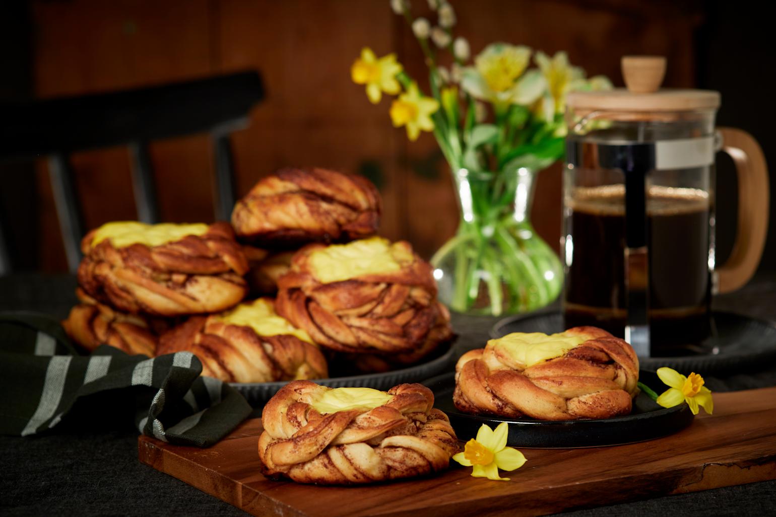 Kanelboller med vaniljekrem servert på både mørk tallerken og mørk trefjøl. Presskanne med kaffe, og vase med påskeliljer i bakgrunnen.