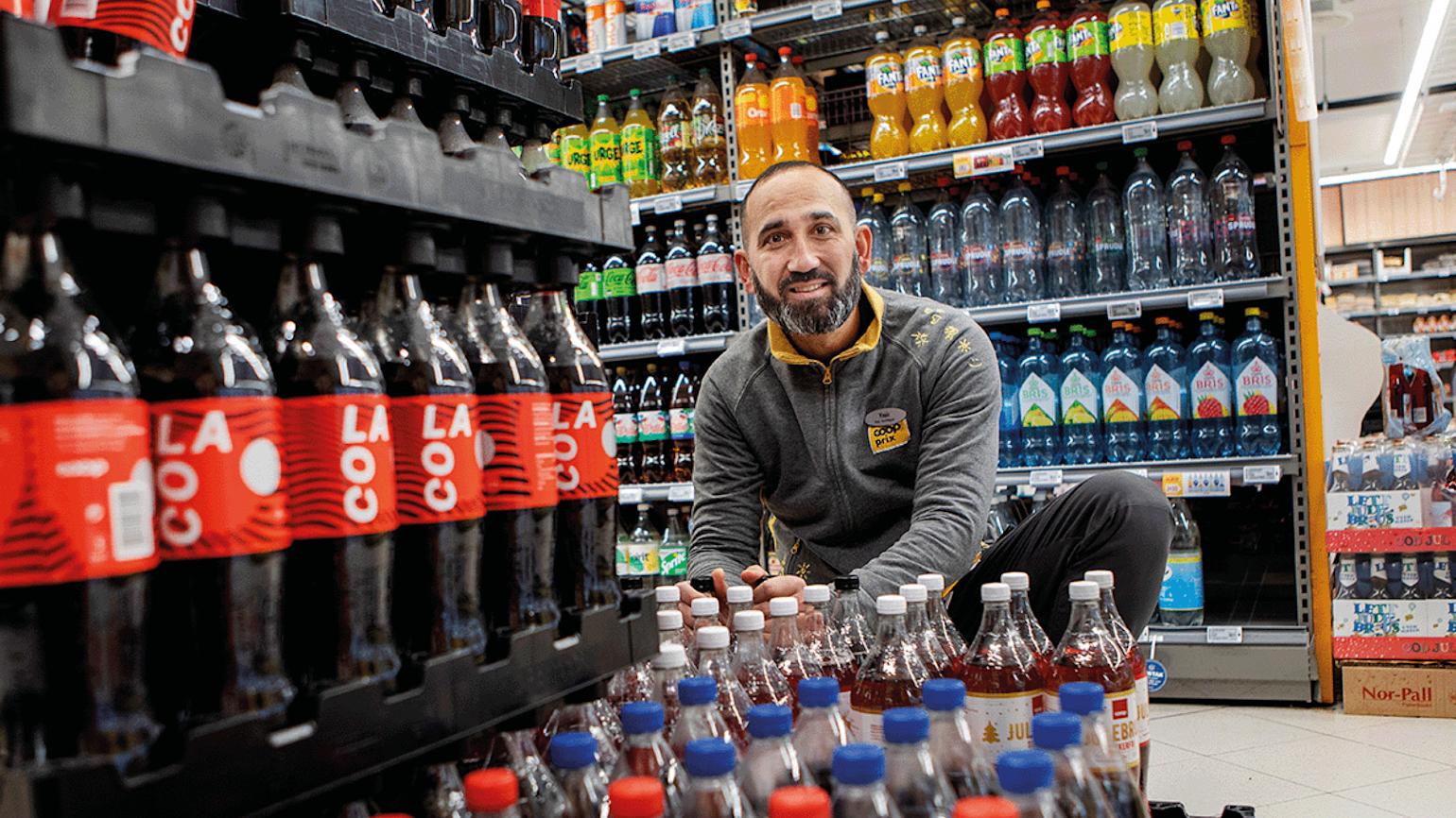 Mann smiler og pakker ut brus i Prix butikk.