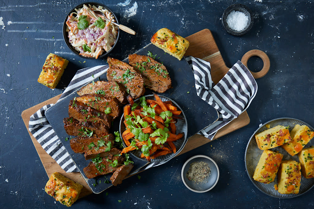 Langtidsgrillet brisket med cheddar jalapeño maisbrød, søtpotetfries og coleslaw servert på steinfjøl på trefjøl med stripete kjøkkenhåndkle. Skåler med tilbehør rundt.