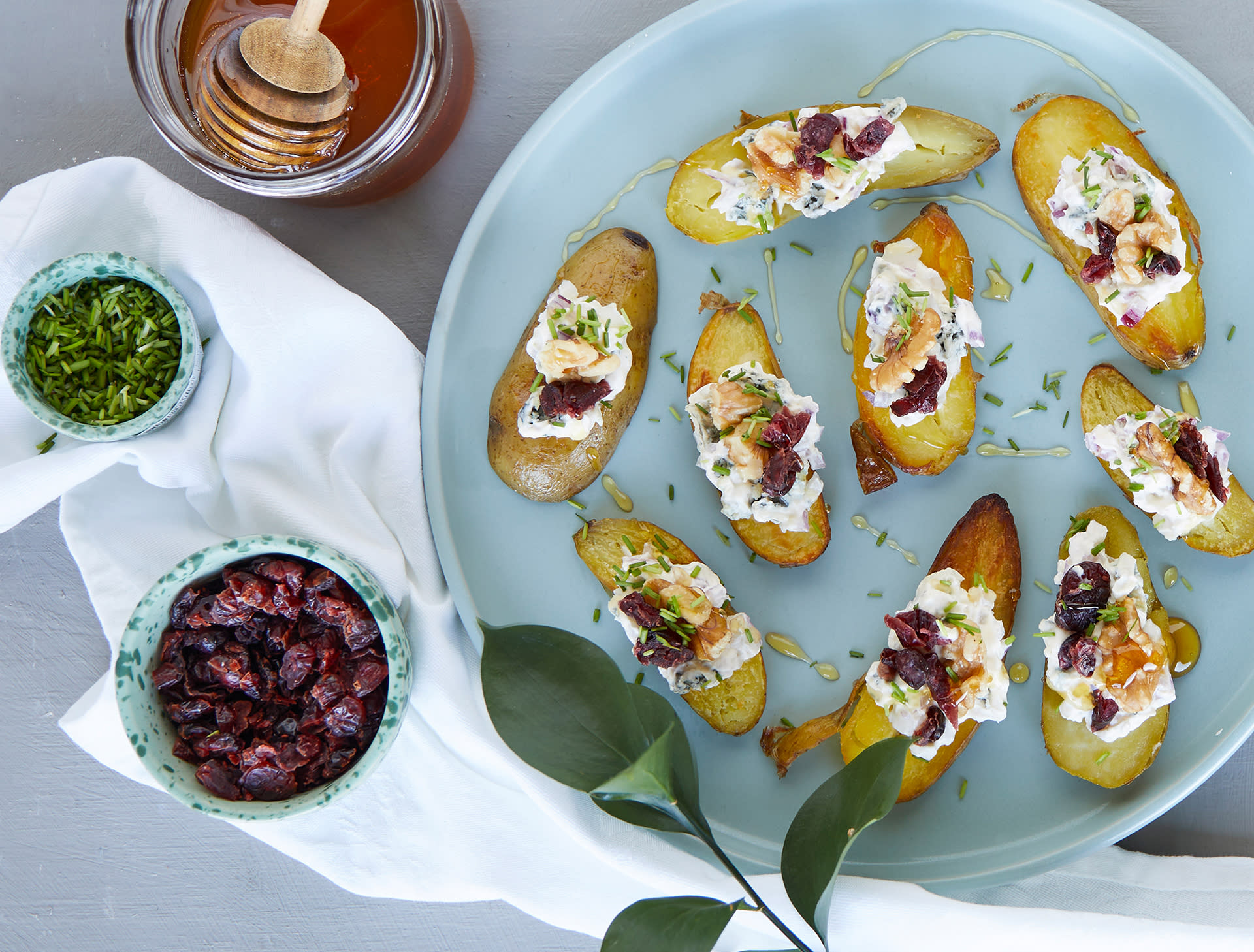 Mandelpotetbruschetta med blåmuggost, tranebær og valnøtter servert på lyst rundt fat med små skåler tørkede tranebær, finhakket gressløk og gyllen honning ved siden av.