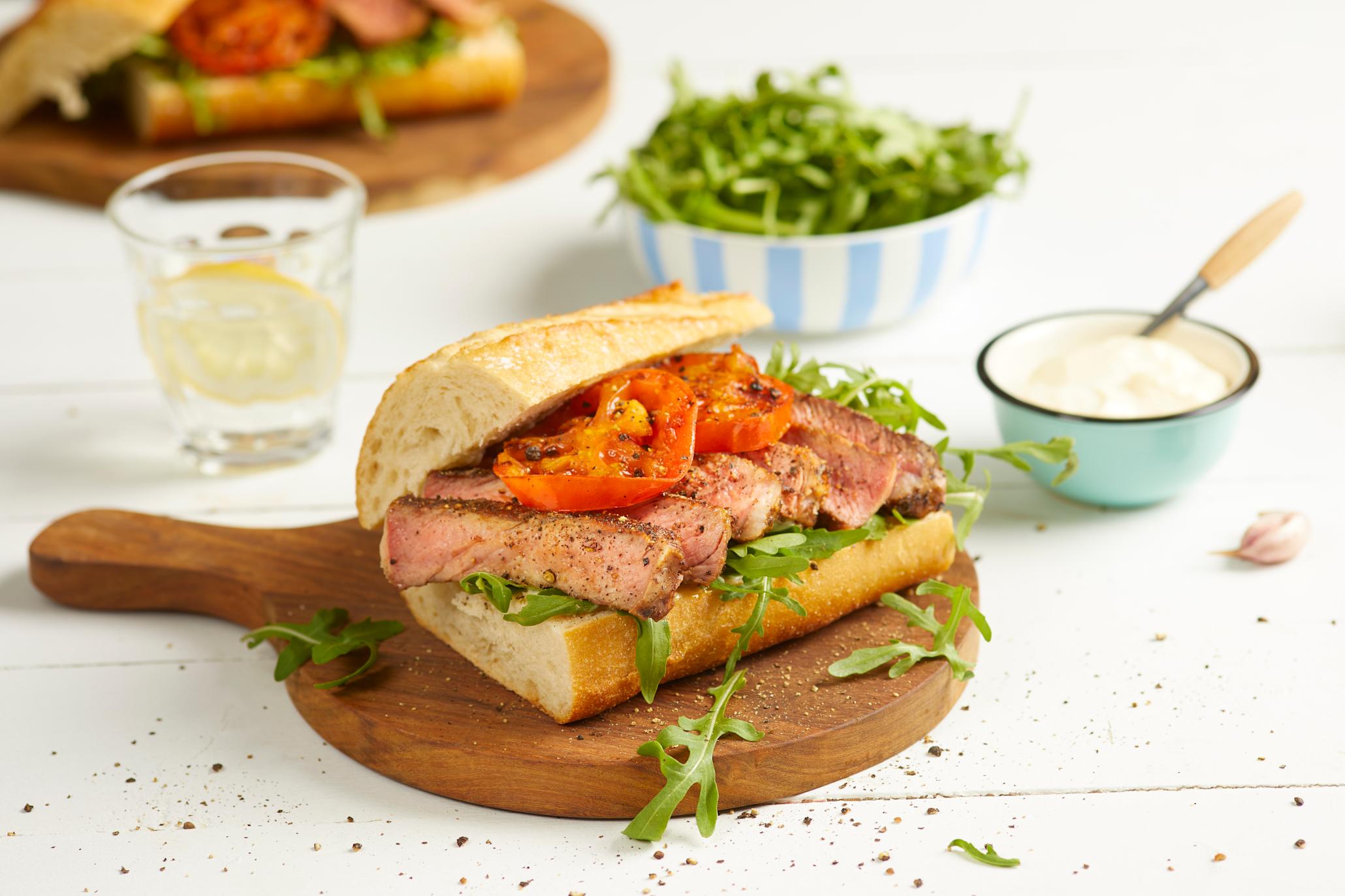 Biffsandwich med grillet tomat og ovnsbakt hvitløksaioli, ruccolasalat og baguette, servert på mørk trefjøl på hvitt trebord. Små skåler med hvitløksaioli og ruccola i bakgrunnen.