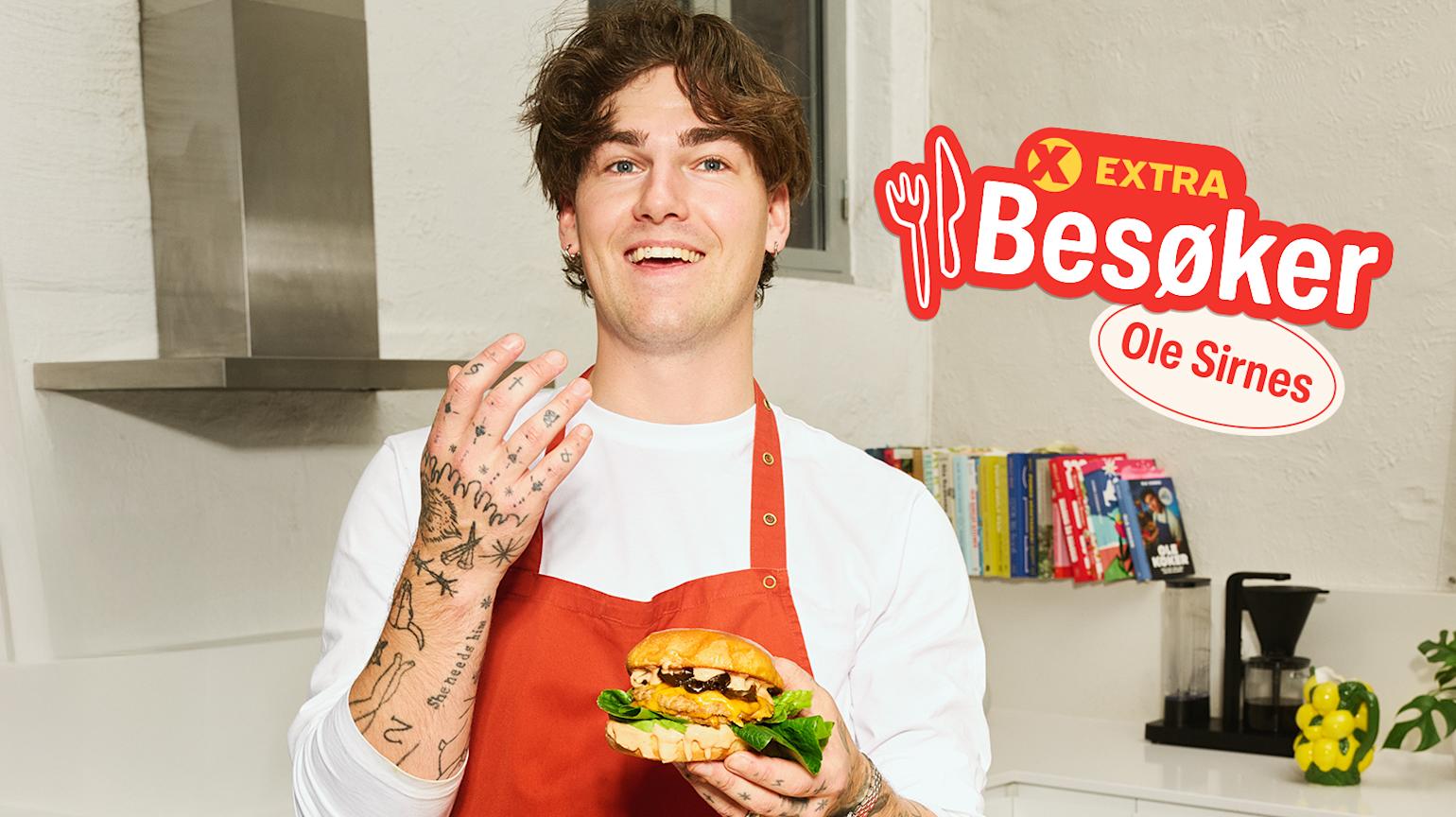 Person i rødt forkle holder en burger på kjøkkenet, med "Extra Besøker Ole Sirnes"-logo i hjørnet.