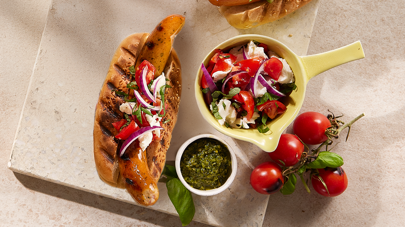 En pølse i brød på et lys marmor fjøl er toppet med tomater, rødløk, mozzarella og basilikum. 4 cherrytomater, en liten hvit skål med pesto og en lysegul skål med tomater, rødløk, mozzarella og basilikum ligger ved siden av pølsen.
