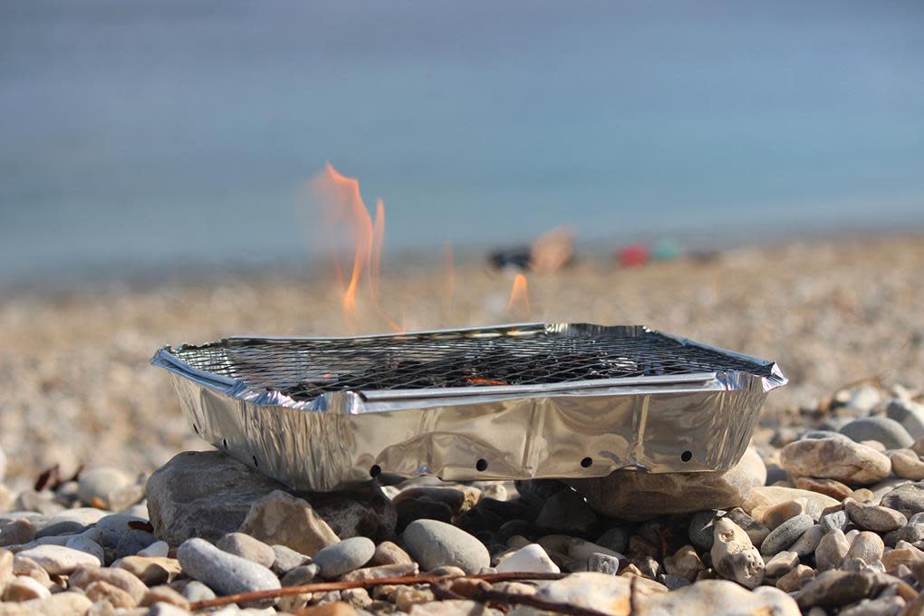 Engangsgrill med flammer, står på steiner på en strand.